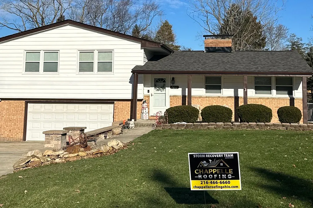 A suburban house with a beige exterior, featuring a garage and a front porch, with a yard sign advertising "Our Work" in roofing services.