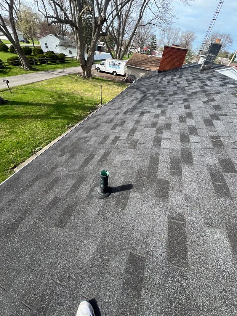 View of a roof with new dark shingles and a vent pipe in the center. This recent work showcases our roofing services. In the background, trees, a grassy yard, and parked vehicles complete the scene.