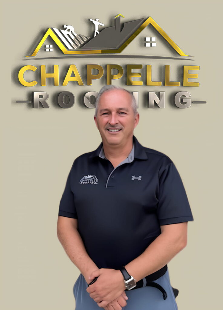 A man with gray hair stands smiling in front of a logo for Chappelle Roofing. He is wearing a navy-blue polo shirt, embodying the professionalism and friendly service that defines our team. Learn more on our About Us page.