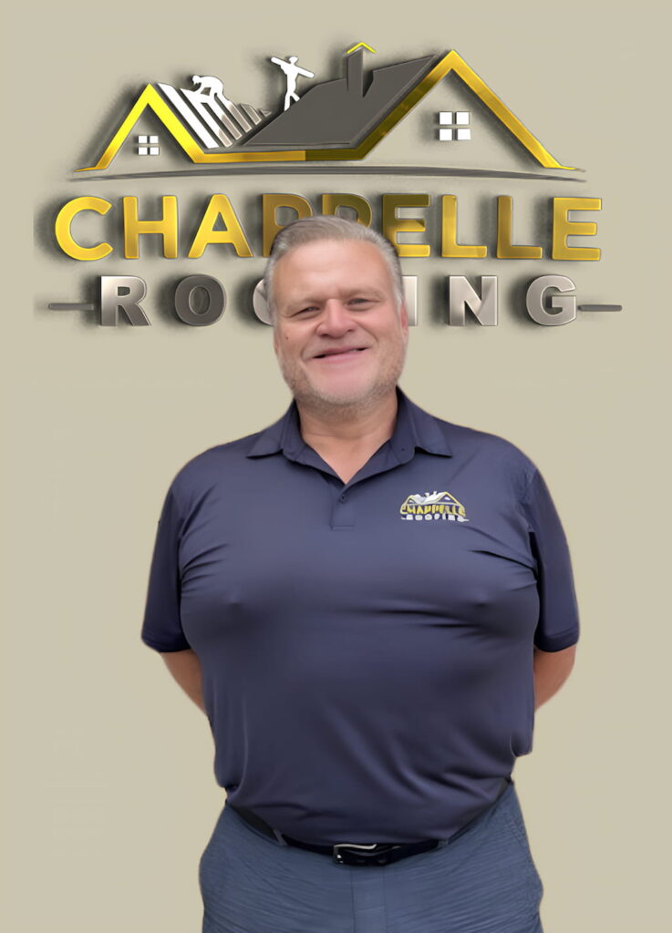 A person is standing and smiling in front of a wall with a "Chappelle Roofing" logo. They are wearing a dark blue shirt that also features the company logo, embodying the spirit of Chappelle Roofing's commitment to quality. Learn more about us through their dedicated service.