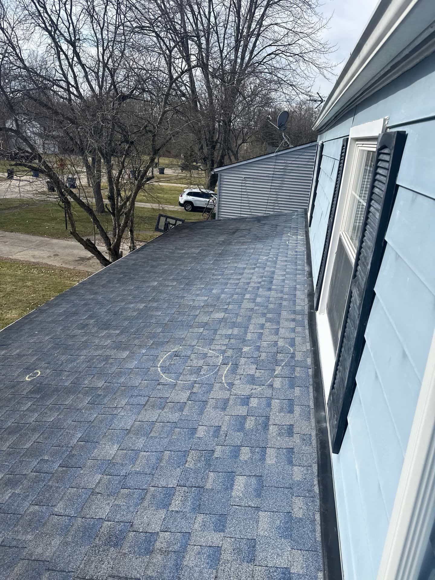 View of a rooftop with blue shingles, part of a light blue house, and bare trees in the background. A white car is parked on a street visible in the distance, showcasing our work's attention to detail and quality.