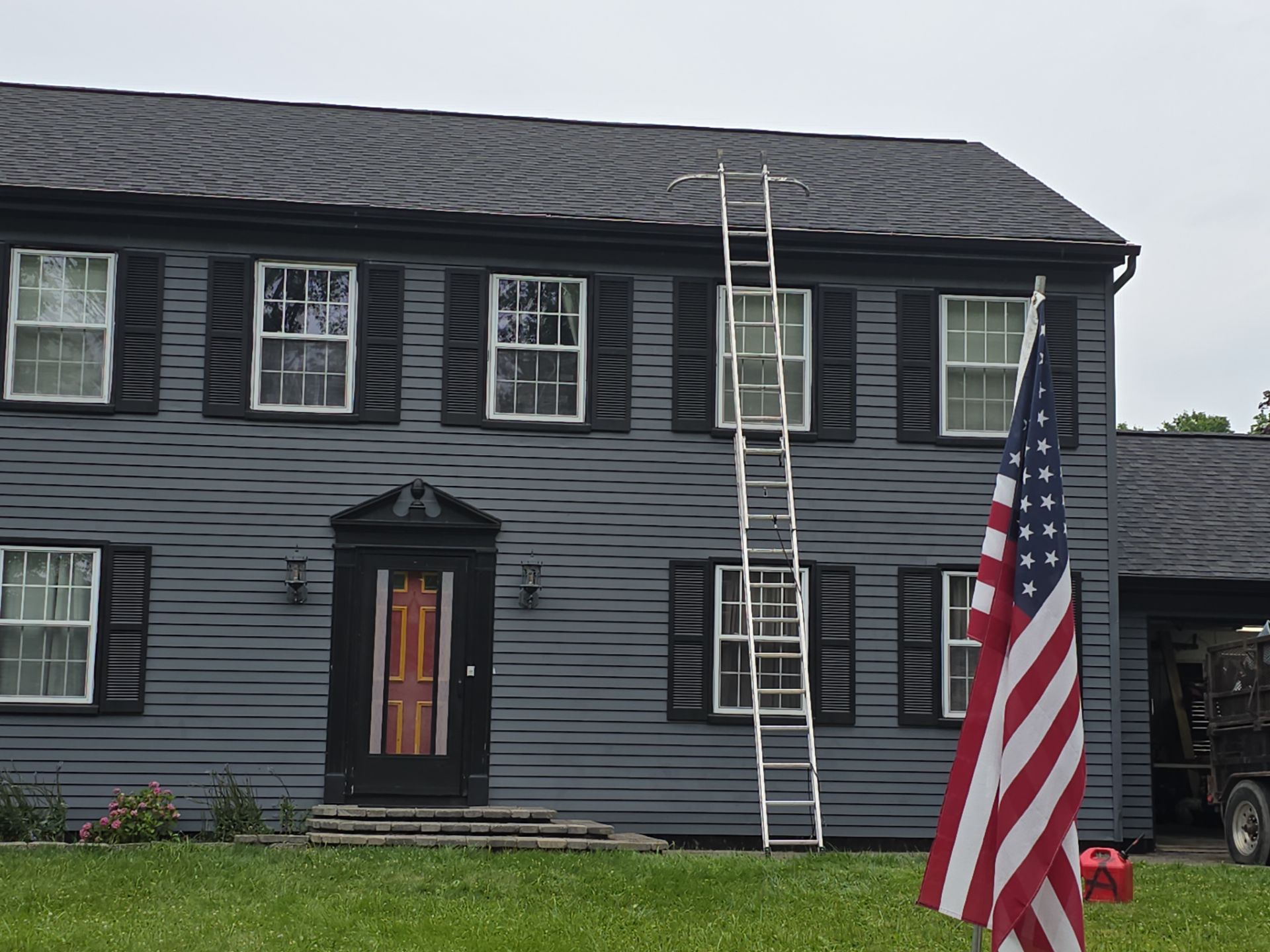 A two-story gray house with black shutters is shown. There is a ladder leaning against the roof on the right side. The U.S. flag is in the foreground on the right.