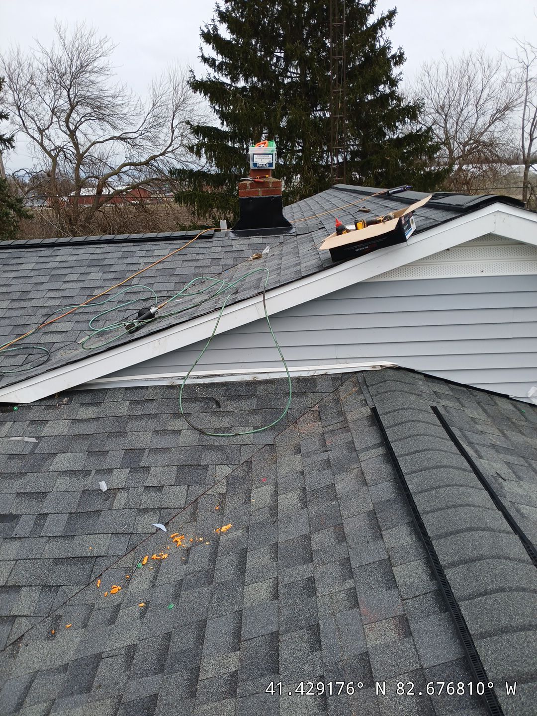 A rooftop with grey shingles features a variety of tools and materials, including wires and wooden planks, scattered around. GPS coordinates are displayed at the bottom. Surrounding area includes bare trees.